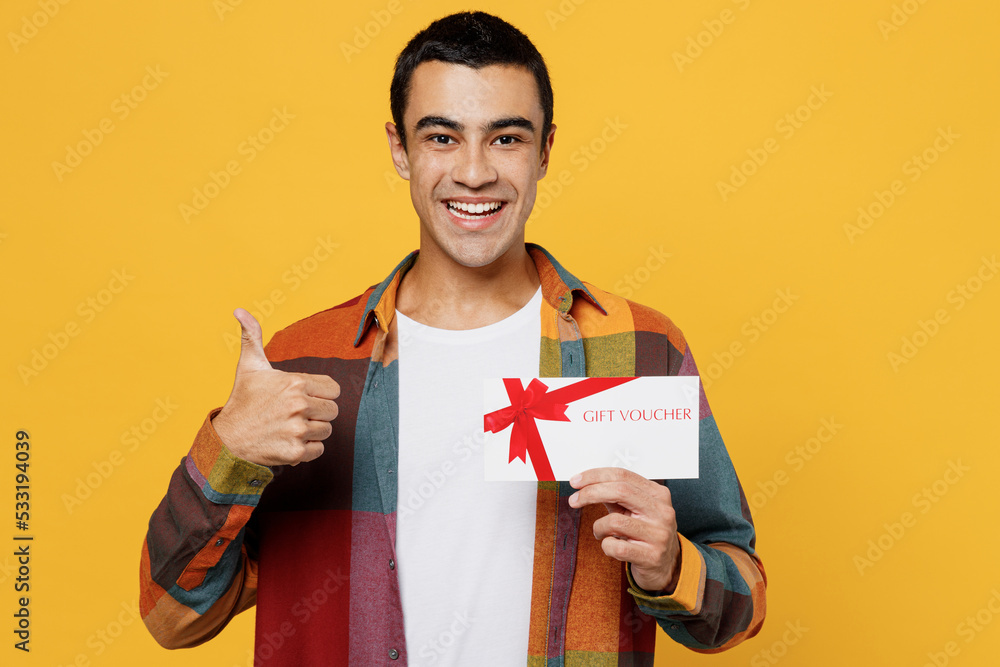 Young happy middle eastern man he wear casual shirt white t-shirt hold gift certificate coupon voucher card for store show thumb up isolated on plain yellow background studio People lifestyle concept.