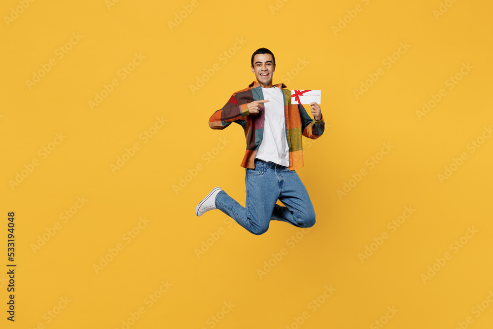 Full body young surprised happy middle eastern man wear casual shirt white t-shirt jump high hold point finger on gift certificate coupon voucher card for store isolated on plain yellow background.