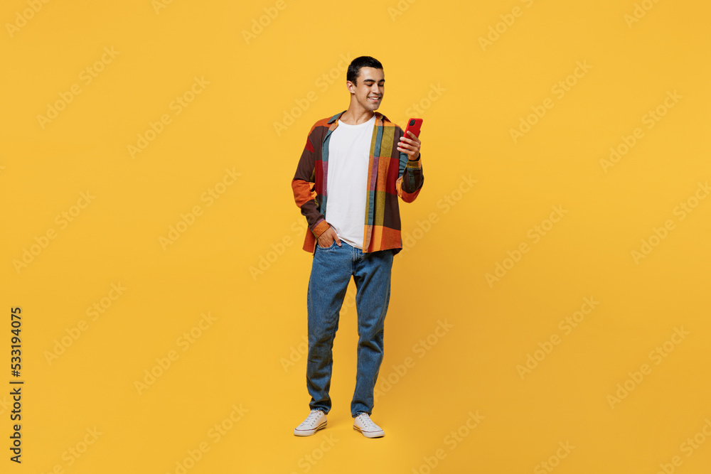 Full body smiling happy fun young middle eastern man he wear casual shirt white t-shirt hold in hand use mobile cell phone isolated on plain yellow background studio portrait People lifestyle concept. Full body smiling happy fun young middle eastern man he wear casual shirt white t-shirt hold in hand use mobile cell phone isolated on plain yellow background studio portrait People lifestyle concept.