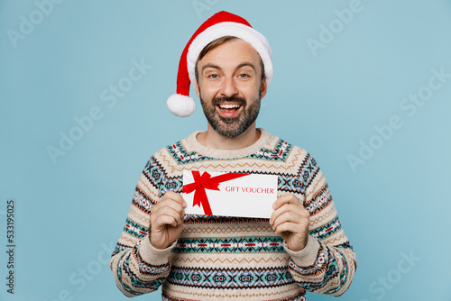 Young man wear sweater christmas Santa Claus hat hold gift certificate coupon voucher card for store isolated on plain pastel light blue cyan background Winter holidays New Year celebration concept.