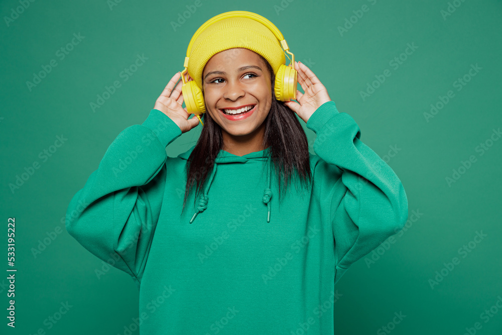 Little happy minded fun kid teen girl of African American ethnicity 13-14 years old wear hoody hat headphones listen music look aside on area isolated on plain dark green background Childhood concept.