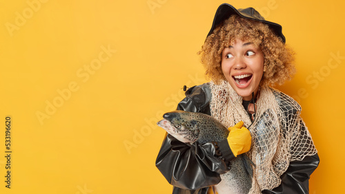 Horizontal shot of happy surprised female angler poses with fish net and stout has good successful day for fishing dressed in leather hat and raincoat isolated over yellow background blank space