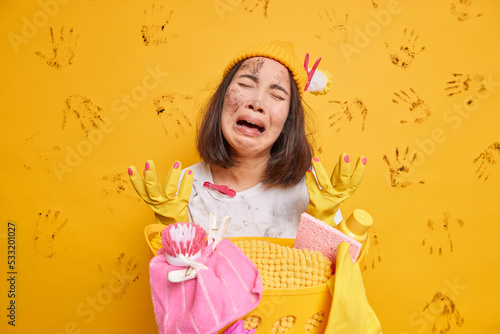 Desperate exhausted Asian woman cries from despair raises hands wears hat and rubber gloves does washing poses near basket of laundry tired of housework isolated over yellow dirty background