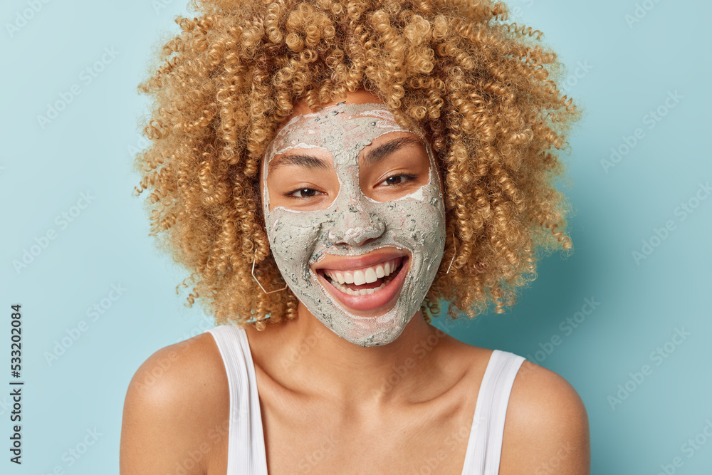 Close up portrait of cheerful curly haired woman smiles broadly applies nourishing clay mask on face to reduce fine lines dressed in t shirt isolated over blue background. Skin care routine concept Close up portrait of cheerful curly haired woman smiles broadly applies nourishing clay mask on face to reduce fine lines dressed in t shirt isolated over blue background. Skin care routine concept