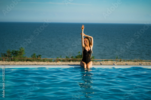 A slender woman in a black swimsuit sits on the side of a panoramic pool. 