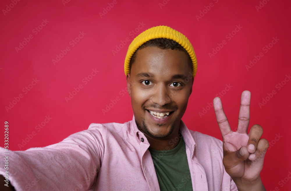 Handsome young african american man, guy making selfie gesturing V, victory or five using smartphone wearing pink shirt and yellow hat isolated on red background Handsome young african american man, guy making selfie gesturing V, victory or five using smartphone wearing pink shirt and yellow hat isolated on red background
