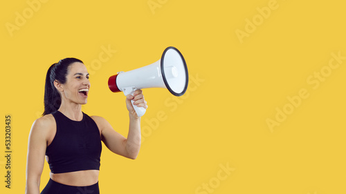 Beautiful fit active woman with megaphone standing on bright yellow copy space background, inviting to fun sports event, announcing discounts at fitness club or promoting great sale at shopping centre