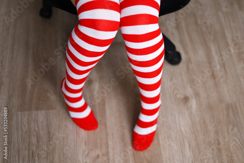 Female legs in Christmas knee socks. Woman sitting in office chair, concept of New Year celebration, girl in Santa costume