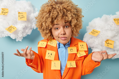 Confused hesitant woman with curly hair wears elegant prange jacket with stuck sticky notes on it has many tasks to do cannot decide from what to start isolated over blue backgound clouds above photo