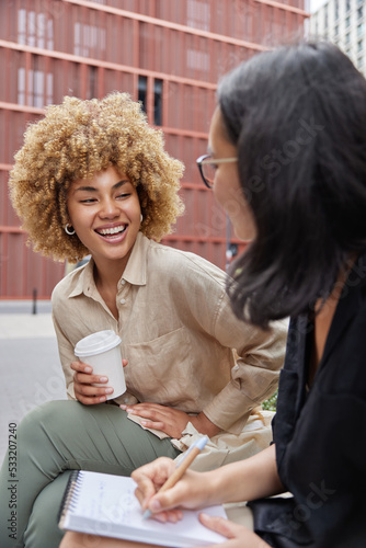 Outdoor shot of two happy diverse female students meet together discuss future project write down information in notebook drink coffee to go laugh joyfully have good mood. Cooperation concept