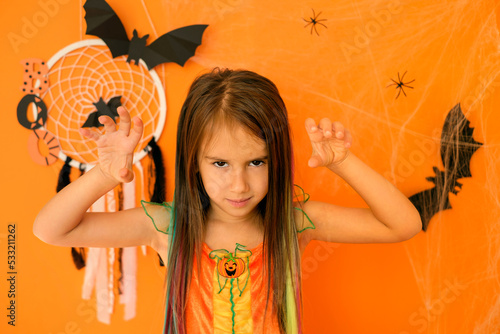 An evil girl intimidating with her formidable appearance in the guise of a witch on an orange background with a Halloween decor with cobwebs and bats
