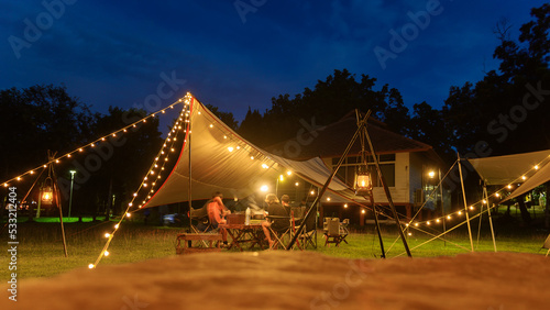 Outdoor camping tent with tarp or flysheet on grass courtyard and warm night light under dark blue sky twilight time, family vacation picnic on holiday relax, Overview of camping of family tourist.