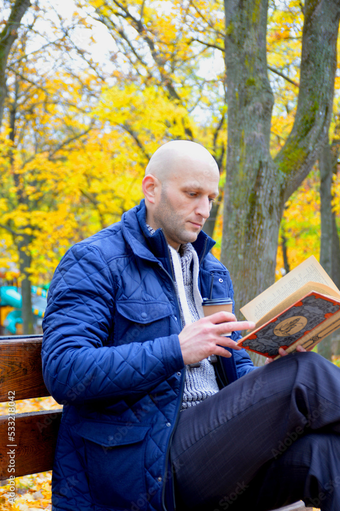 Fototapeta premium young man reads a book and drinks coffee outdoors, sitting on a bench in an autumn park. serenity, relaxation and privacy concept