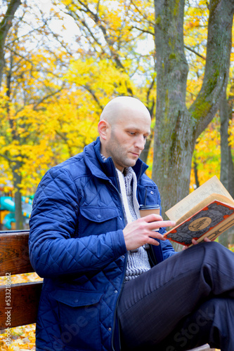 young man reads a book and drinks coffee outdoors, sitting on a bench in an autumn park. serenity, relaxation and privacy concept