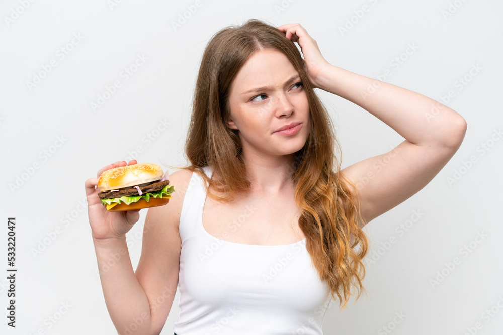 Young pretty woman holding a burger isolated on white background having doubts and with confuse face expression Young pretty woman holding a burger isolated on white background having doubts and with confuse face expression