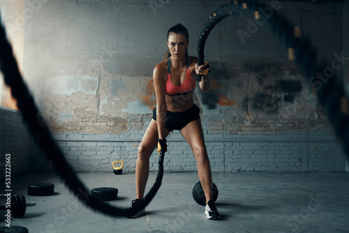Full length of beautiful young woman exercising with battle ropes in gym
