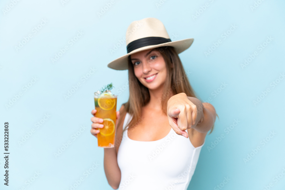 Young beautiful woman holding a cocktail isolated on blue background points finger at you with a confident expression Young beautiful woman holding a cocktail isolated on blue background points finger at you with a confident expression