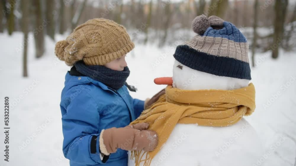 Little boy building snowman in snowy park. Child puts hat and scarf on snowman . Active outdoors leisure with family with children in winter. Kid during stroll in a snowy winter park