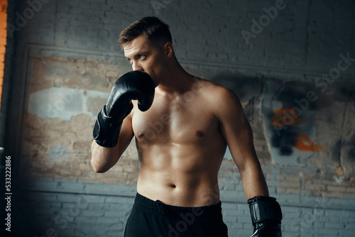 Confident young man in boxing gloves touching face and looking away while standing in gym