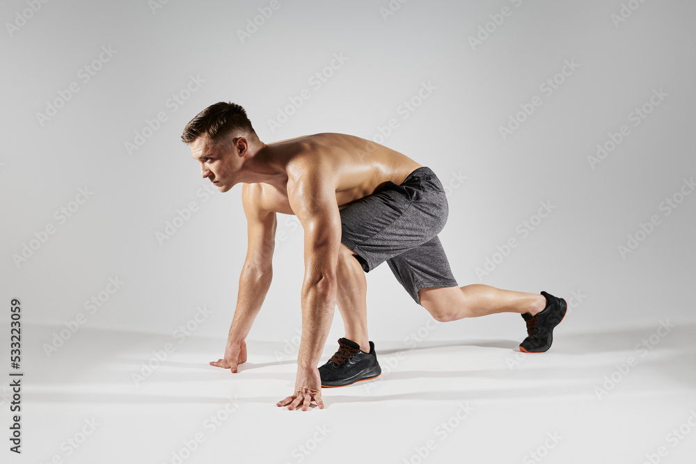 Young fit man preparing to run while standing in starting position against white background - obrazy, fototapety, plakaty 