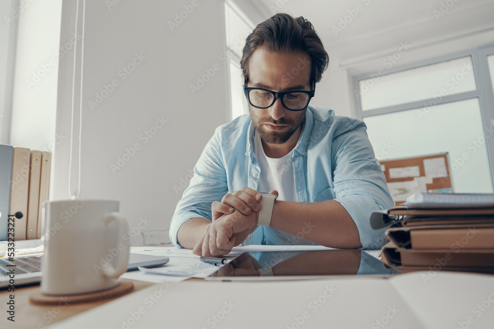 Handsome young man looking at his smart watch while sitting at his working place in office Handsome young man looking at his smart watch while sitting at his working place in office