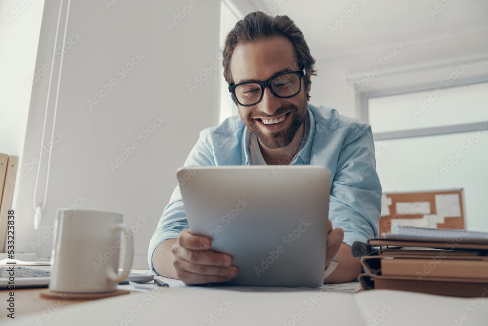 Handsome young man working on digital tablet and smiling while sitting at his working place in office Handsome young man working on digital tablet and smiling while sitting at his working place in office