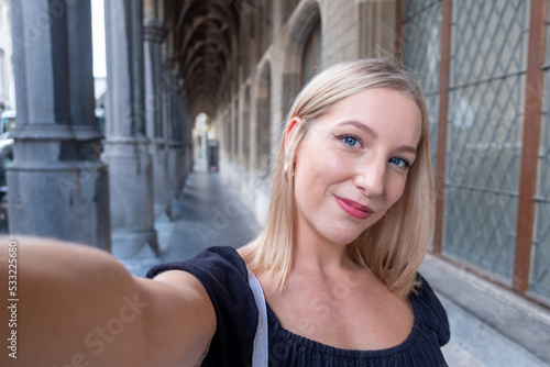 Portrait of wonderful white female model with bright makeup expressing energy in good day in Europe. Lovely blonde woman in stylish clothes making selfie while walking past old building. High quality