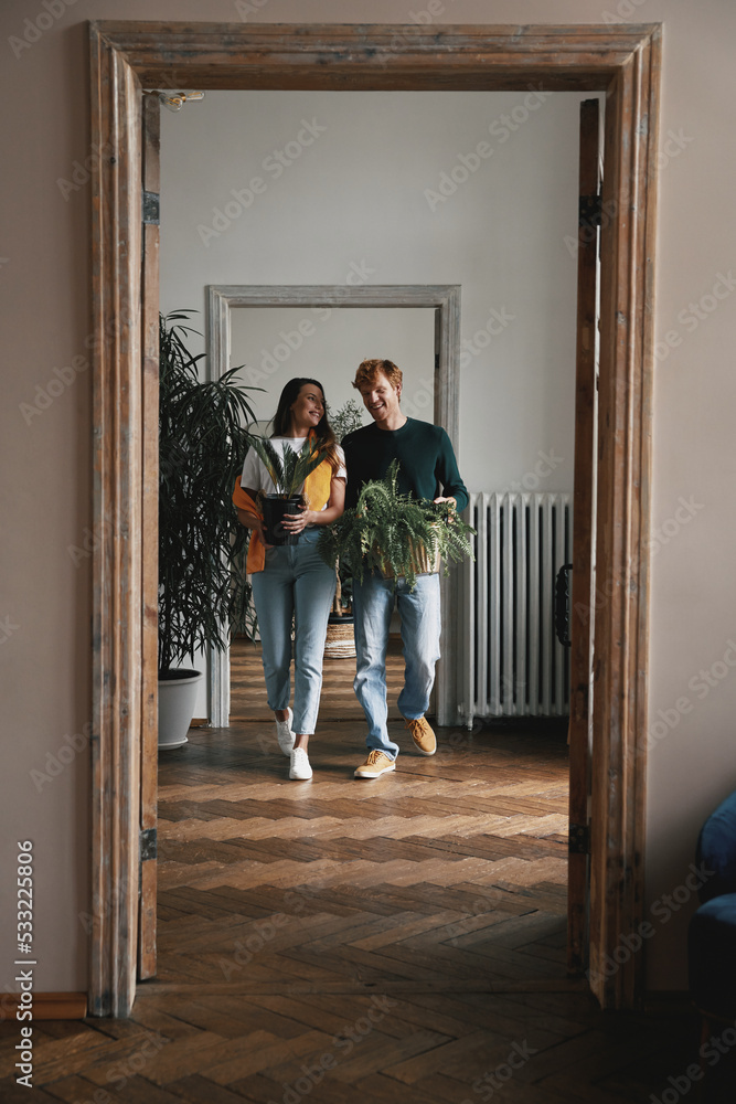 Happy young couple holding houseplants while walking by their new apartment together - obrazy, fototapety, plakaty 