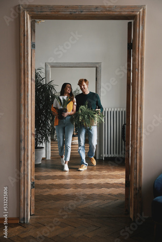 Happy young couple holding houseplants while walking by their new apartment together © gstockstudio