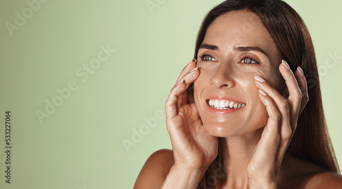 Smiling female aging model, clear glowing skin, nourished face without blemishes, applies daily cream, hyaluron anti-aging treatment, looking in mirror at herself, green background photo