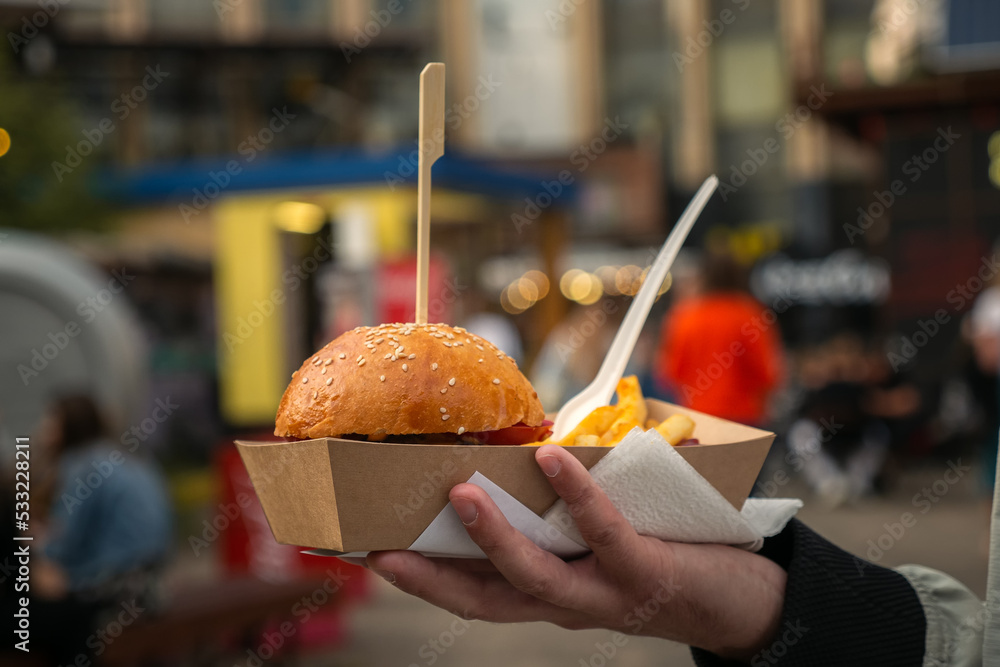 Man hands holding street food burger with french fries on craft paper. Street fast food. festival background