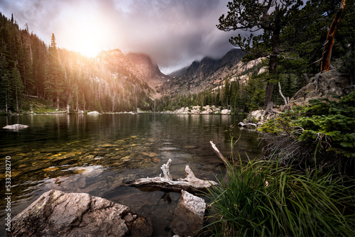 Mountain lake at sunset with granite rock and trees along the shoreline. Orange sunlight is shining down onto the lake from the top of a mountain peak. 