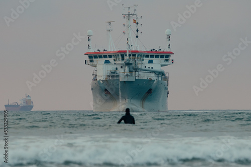 surfers in the water in the ocean waves while watching a large industrial ship in the middle of the sea during a surf and travel week experience in Somo, Cantabria (Spain)