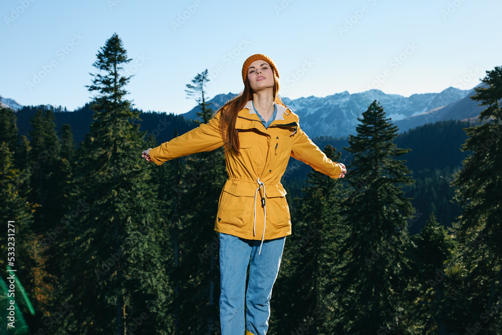 Woman with red hair hiker standing on the mountain hands up happiness overlooking snowy mountains and trees in yellow raincoat and cap travel autumn and hiking in the mountains in the sunset freedom