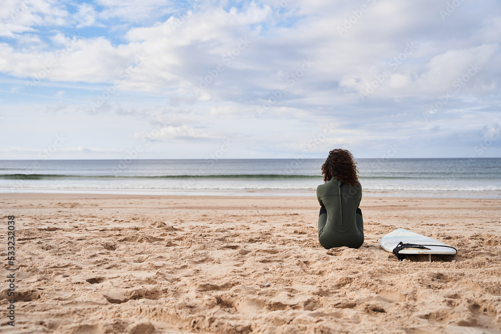 Unrecognizable young surfer woman sitting at the sand with surfboard