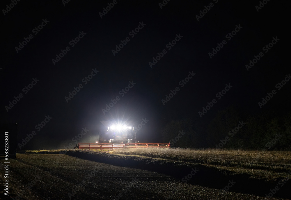 Combine harvester working at night with front headlights illuminating the field during the last days of harvesting season