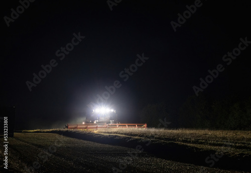 Combine harvester working at night with front headlights illuminating the field during the last days of harvesting season