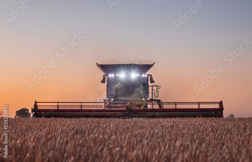 Obraz na płótnie Agriculture: Combine harvester with headlight illuminating wheat field as evening turns into night w salonie