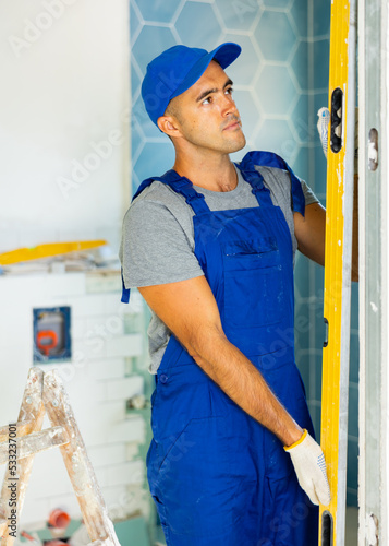 Caucasian repairman in overall measuring wall with spirit level during home improvement works.
