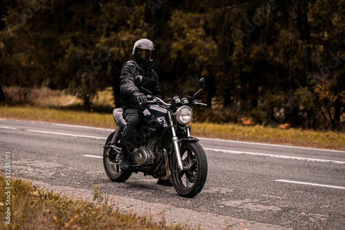 motorcyclist in a motorcycle jacket and a helmet with a sun visor on a custom motorcycle cafe racer. Stylish motorcyclist and autumn road. © velimir