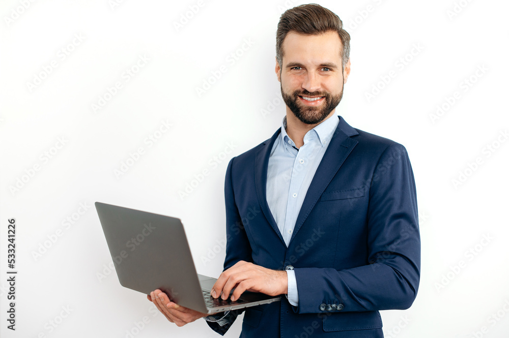 Friendly attractive caucasian bearded business man, in a suit, programmer, IT specialist, holding opening laptop in hands, standing on isolated white background, looks at camera, smiling Friendly attractive caucasian bearded business man, in a suit, programmer, IT specialist, holding opening laptop in hands, standing on isolated white background, looks at camera, smiling