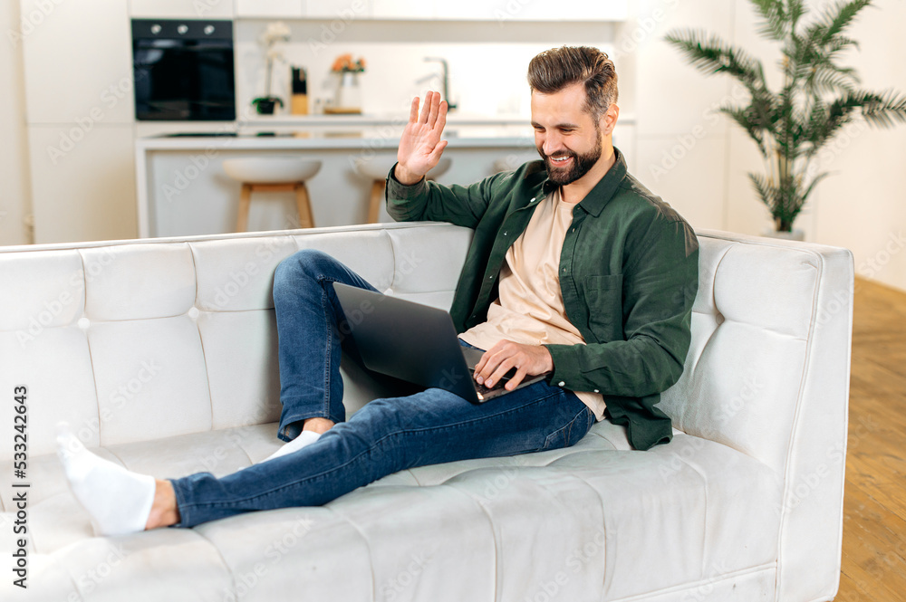 Positive handsome caucasian man, in stylish casual wear, having online video conference with friends or colleagues by laptop at home while sitting on comfortable sofa, waves hand, smiling friendly Positive handsome caucasian man, in stylish casual wear, having online video conference with friends or colleagues by laptop at home while sitting on comfortable sofa, waves hand, smiling friendly