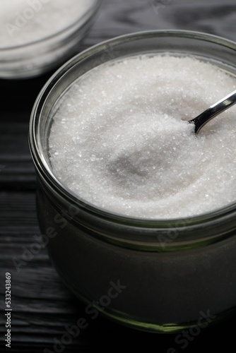 Granulated sugar and spoon in glass jar on black wooden table, closeup