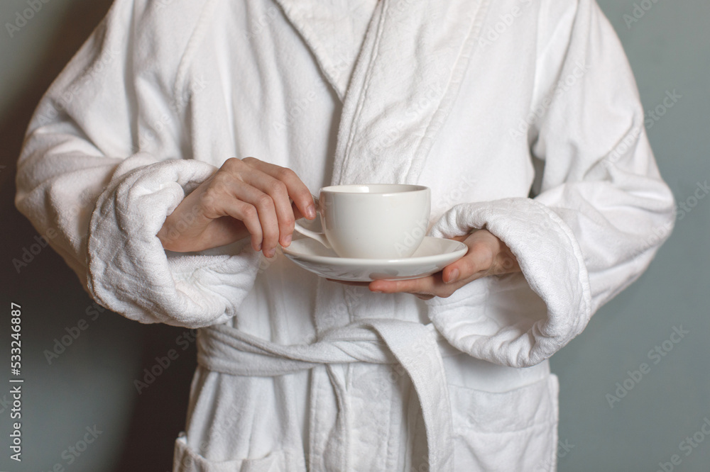 unrecognizable person in white bathrobe holding cup of coffee near gray wall unrecognizable person in white bathrobe holding cup of coffee near gray wall