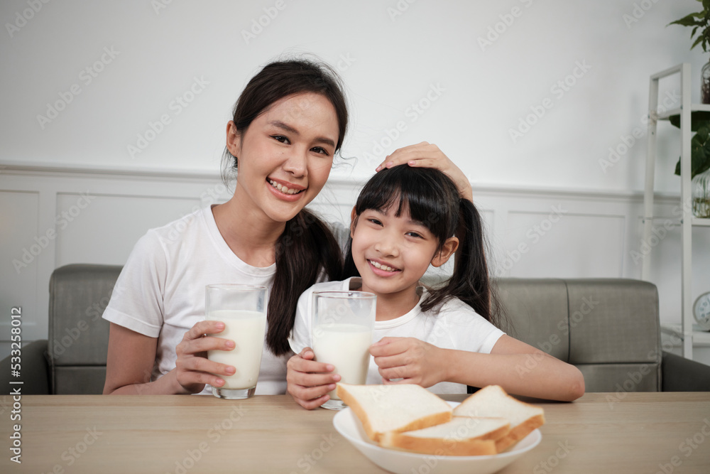 Portrait of healthy Asian Thai family, happy daughter, and young mother looking at camera, drink fresh milk and bread together at dining table in morning, wellness nutrition breakfast meal lifestyle.