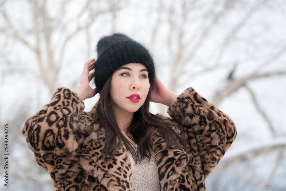 Winter portrait of young adult female with long hair and red plump lips, dressed in fur coat with leopard pattern and black knit hat. Brunette woman raised hands and straightens hat, looking away Winter portrait of young adult female with long hair and red plump lips, dressed in fur coat with leopard pattern and black knit hat. Brunette woman raised hands and straightens hat, looking away