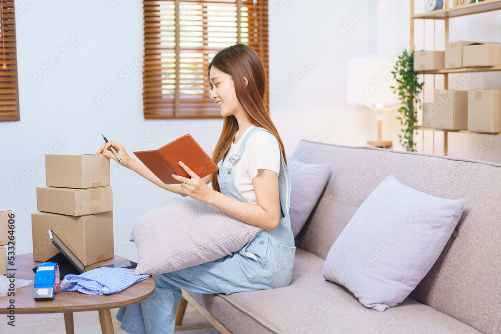 Online trader concept, Female entrepreneur writes on parcel box after reading address on notebook