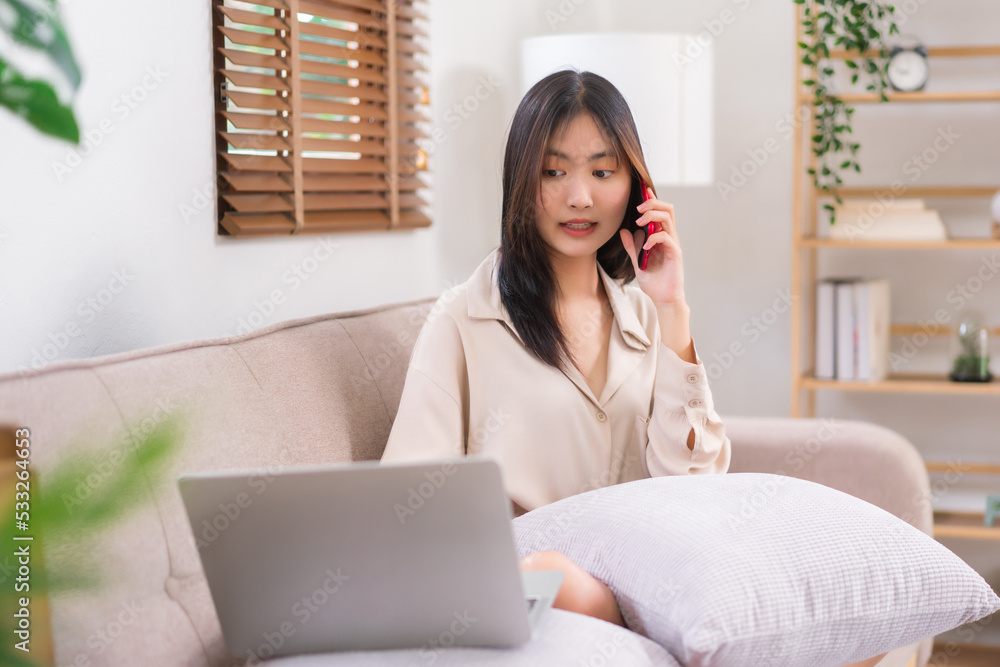 Concept of activity in living room, Young Asian woman use laptop and talking on smartphone on sofa