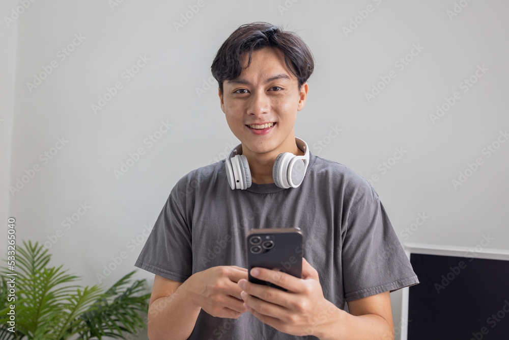 Fototapeta premium Smiling young adult Asian freelance business man using mobile phone checking social media network feed or message chat sitting at home. Online digital communication, rest after hard work day