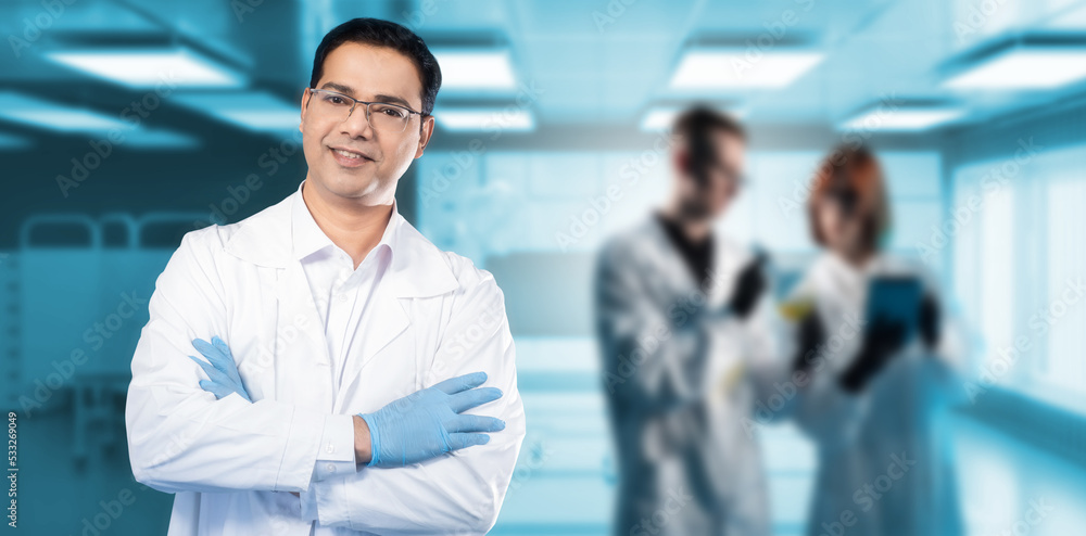 Young scientist. Man doctor in white coat. Scientist stands with arms folded. Man smiles and looks into camera. Confident doctor in clinic. Hospital worker smiles. Doctors in background are blurred Young scientist. Man doctor in white coat. Scientist stands with arms folded. Man smiles and looks into camera. Confident doctor in clinic. Hospital worker smiles. Doctors in background are blurred